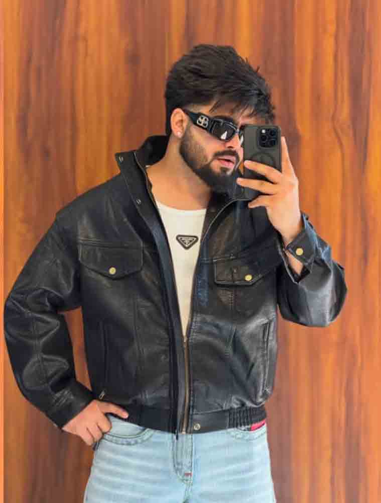 A man taking a mirror selfie wearing a black leather jacket, a white t-shirt with a Prada logo, and light blue jeans against a wooden wall background.