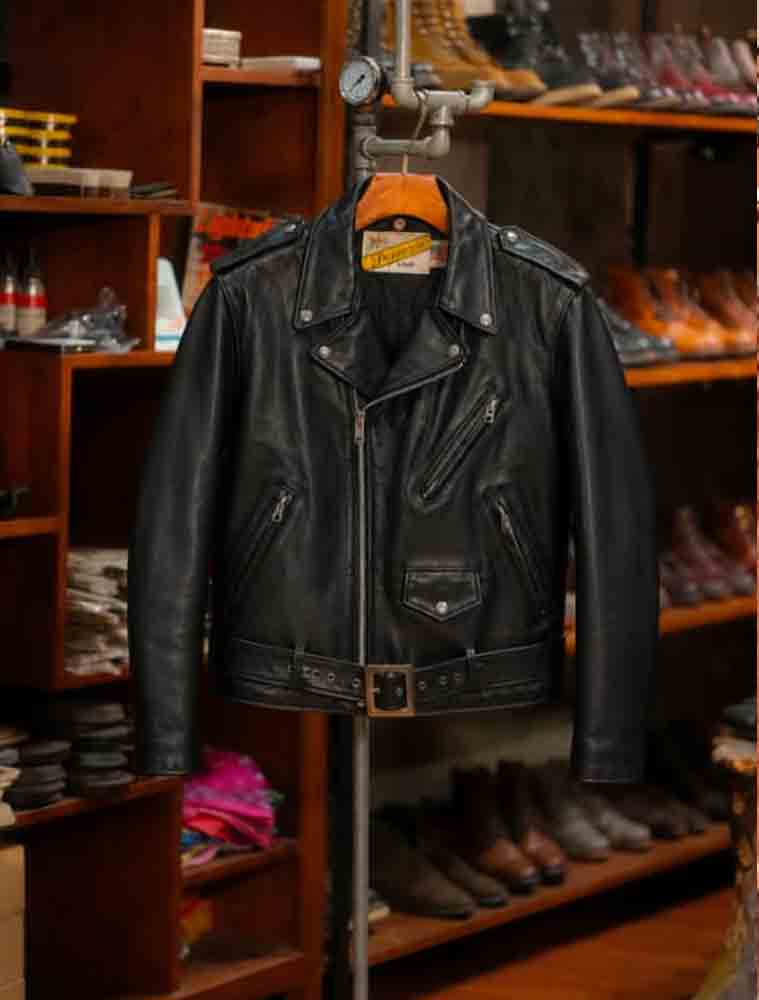 A black leather motorcycle jacket hanging on a wooden hanger on a display stand inside a shoe and leather goods store, with wooden shelves filled with boots and shoes in the background.