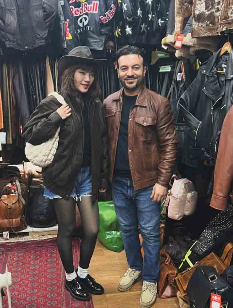 A man and a woman posing together inside a clothing store. The woman is wearing a black wide-brim hat, a black leather jacket, denim shorts, black tights, and black loafers, while carrying a white woven shoulder bag.