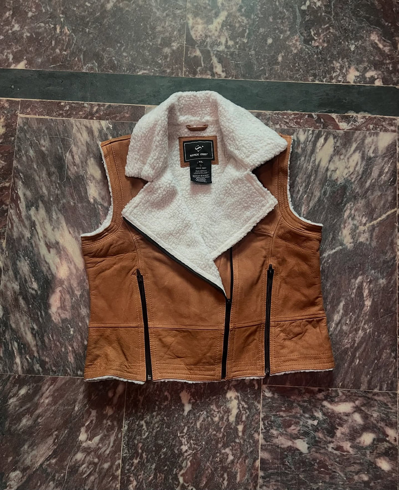 A brown suede vest with a white sherpa lining, laid flat on a dark marble floor. The vest features a wide collar, front zipper closure, and zippered pockets.