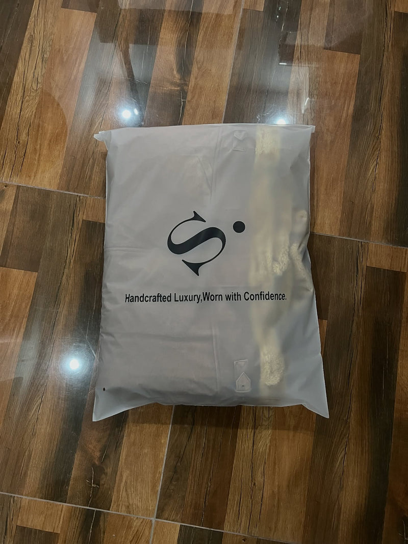 A light gray fabric bag or pouch with a black stylized logo and the text 'Handcrafted Luxury. Worn with Confidence' placed on a brown wood-look tiled floor.