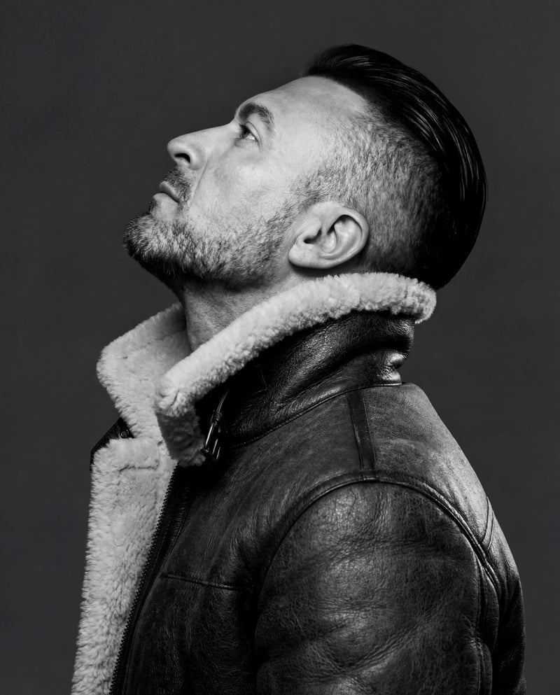 A black and white portrait of a man wearing a dark leather aviator jacket with a thick shearling-style collar, looking upward against a dark gray background.