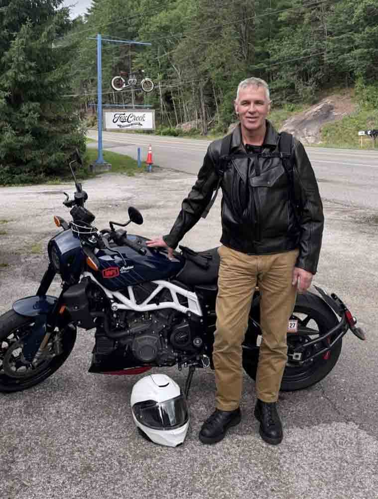 A man in a black leather motorcycle jacket and tan pants stands next to a black and white cruiser motorcycle on a paved road, with a white helmet on the ground in front of it. Trees and a road sign are visible in the background.