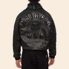 Load image into Gallery viewer, Black leather jacket with 'Avirex USA' branding on the back against a beige background

