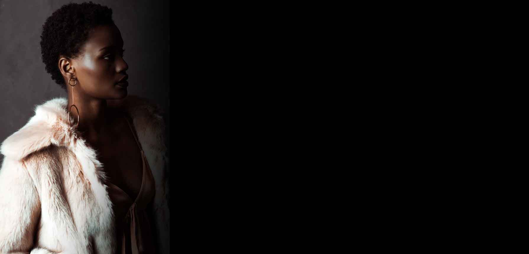 A model in profile wearing a light pink faux fur coat over a brown dress, set against a dark gray background with a large black block on the right side.