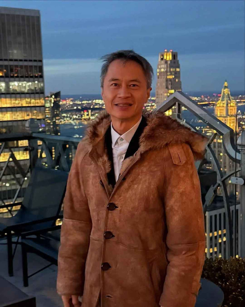 A man wearing a brown shearling-lined coat with a fur-trimmed collar, standing on a rooftop terrace with a city skyline at dusk in the background.