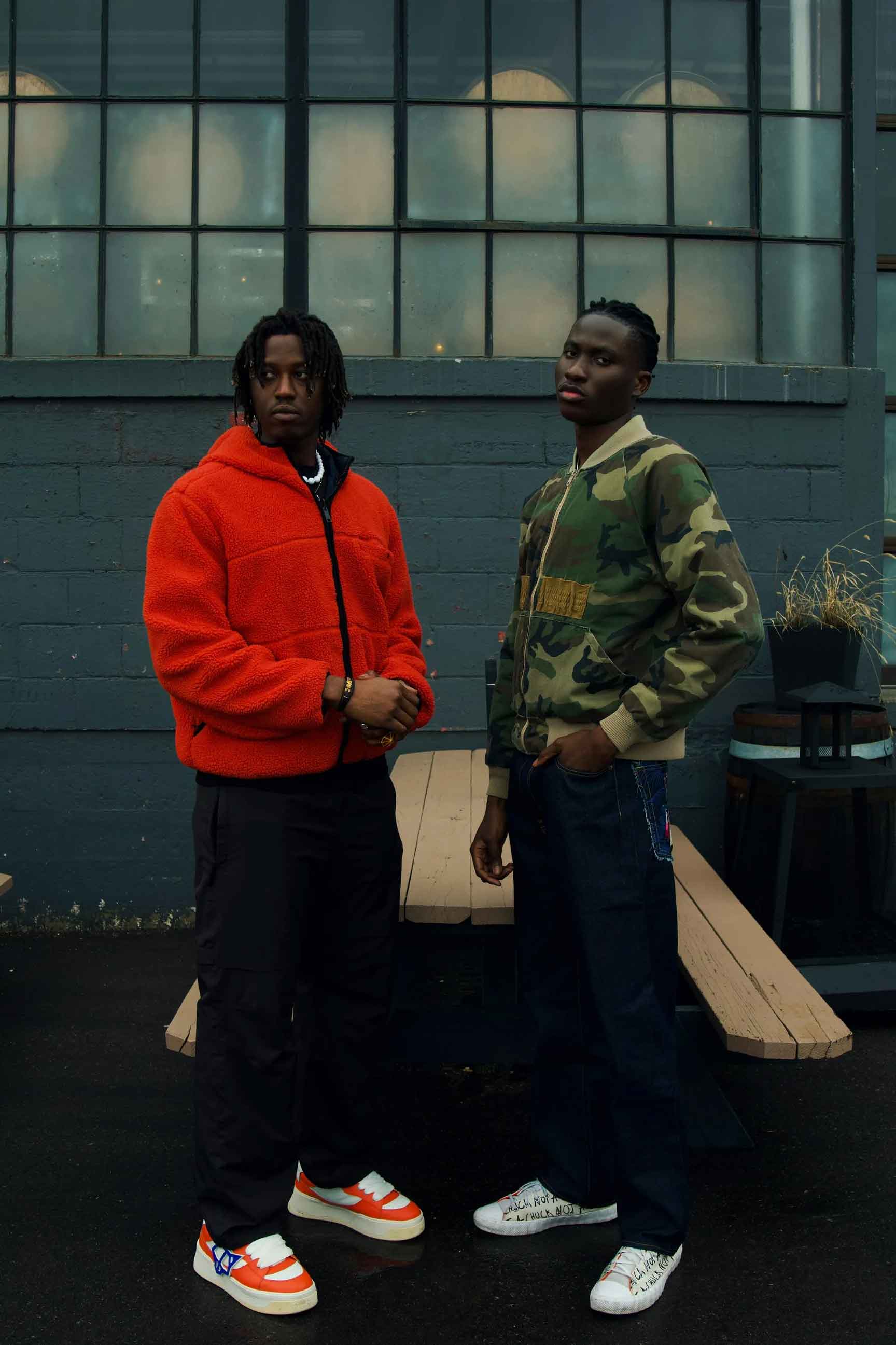 Two men standing outdoors in front of a dark brick wall with a large window. The man on the left wears a bright orange zip-up hoodie, black pants, and orange-and-white sneakers.