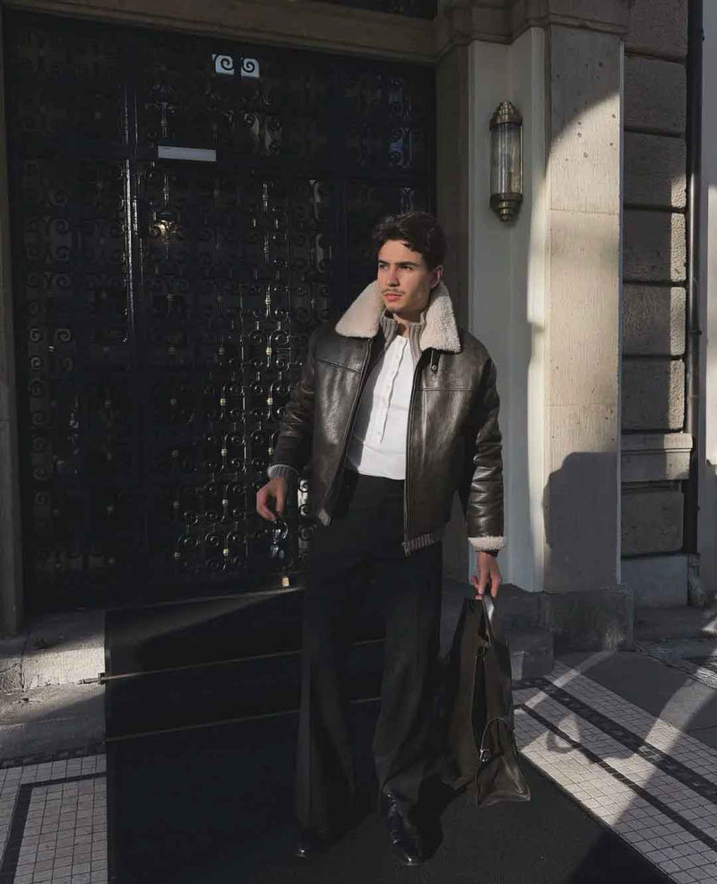 A man wearing a dark brown leather jacket with a cream shearling collar, a white shirt, and dark trousers walks out of a building with strong sunlight and shadows.
