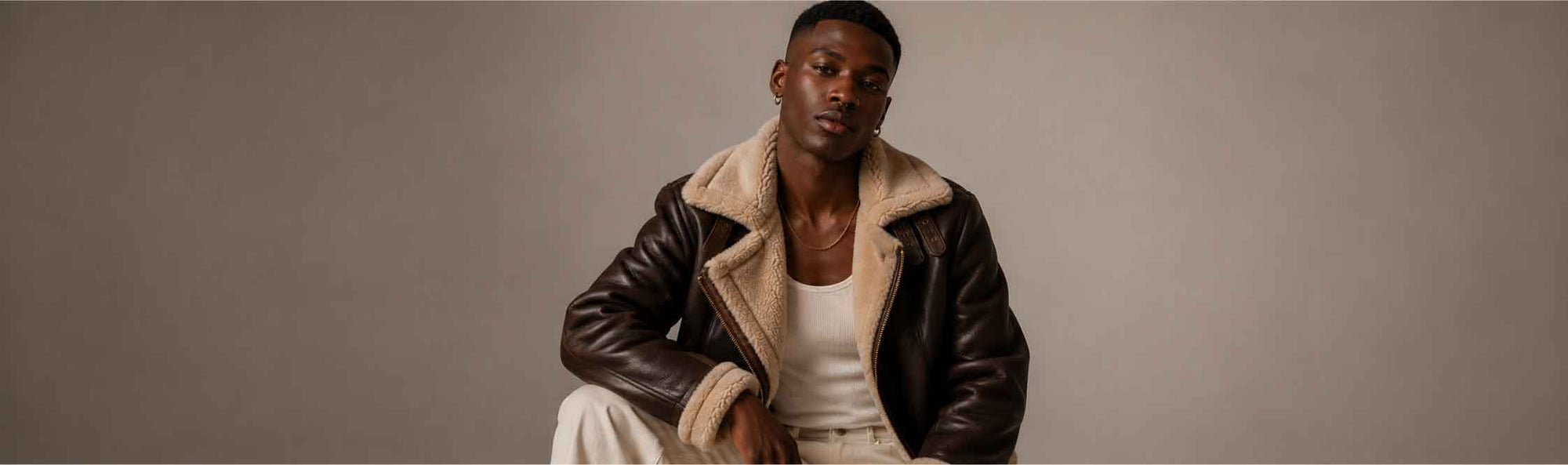 A man wearing a dark brown leather aviator jacket with a beige shearling lining, layered over a white tank top and white pants, seated against a neutral gray studio background.