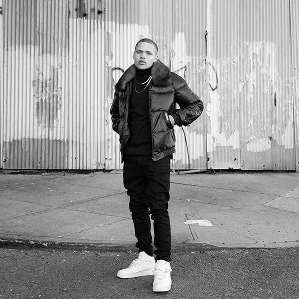 A man wearing a black puffer jacket with a fur collar, black turtleneck, black cargo pants, and white sneakers, standing in front of a corrugated metal wall.