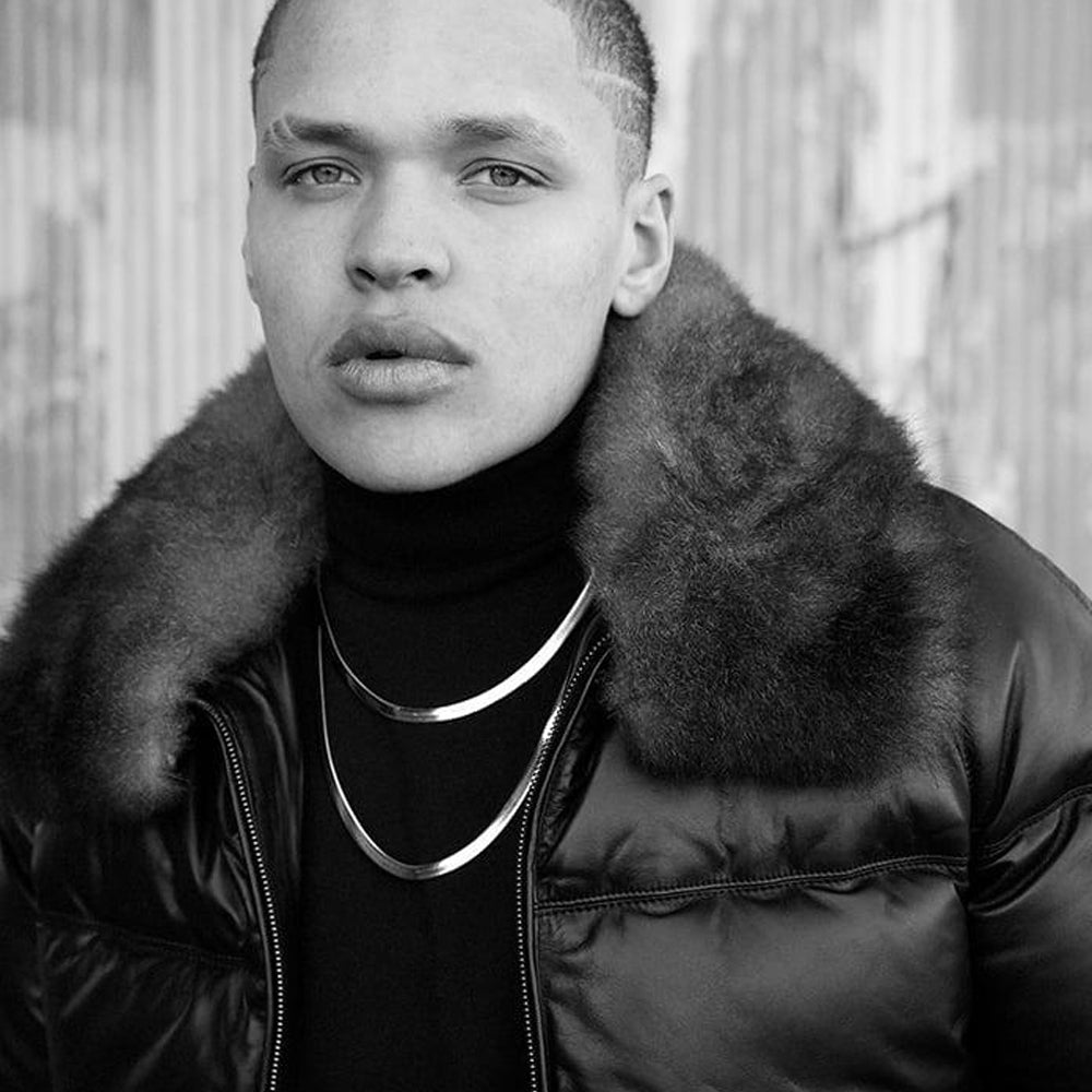 A black and white portrait of a man wearing a black puffer jacket with a large fur collar, layered over a black turtleneck and silver chain necklaces.
