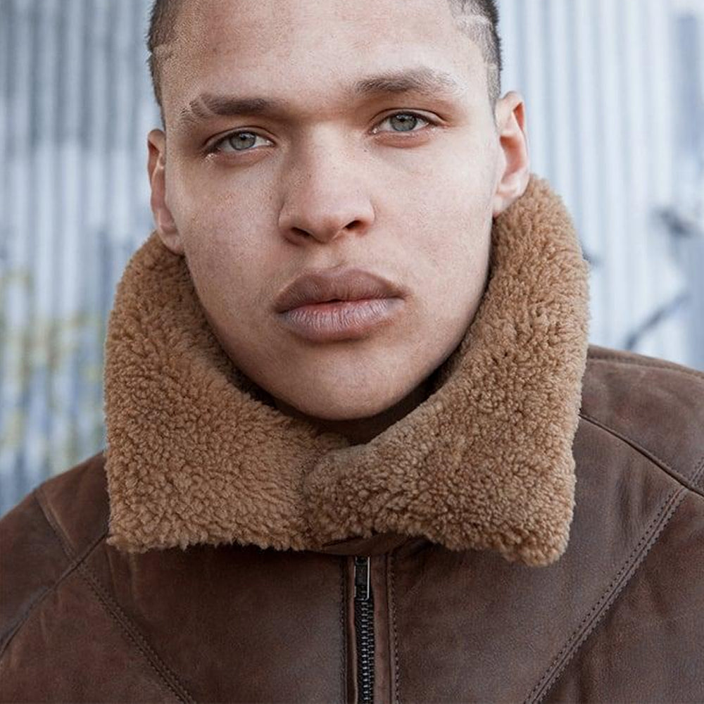 Close-up of a person wearing a brown leather jacket with a large tan sherpa fleece collar, photographed against a softly blurred outdoor background.