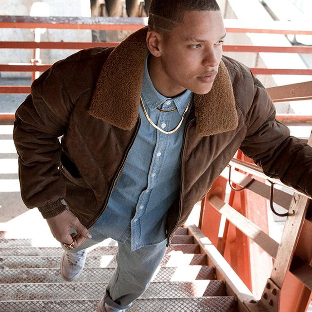 A man wearing a brown suede bomber jacket with a brown sherpa collar, layered over a light blue button-down shirt and jeans, walking down metal stairs.