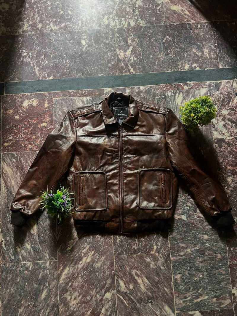 A brown leather jacket with a front zipper and two large patch pockets, laid flat on a dark marble-patterned tile floor with two small green plants as props.