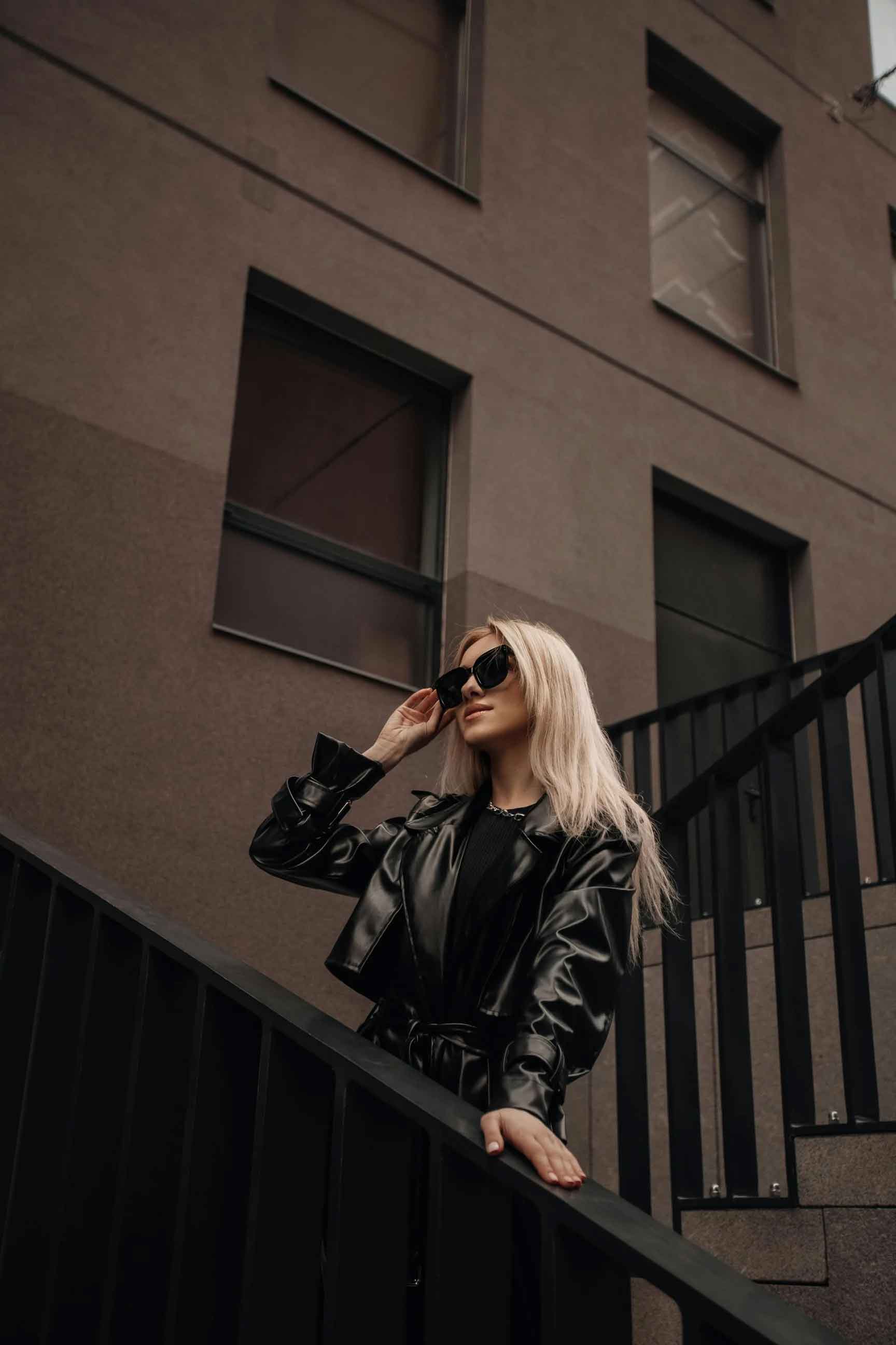 A woman with long blonde hair wearing a shiny black leather jacket and black sunglasses stands on an outdoor staircase in front of a brown building.