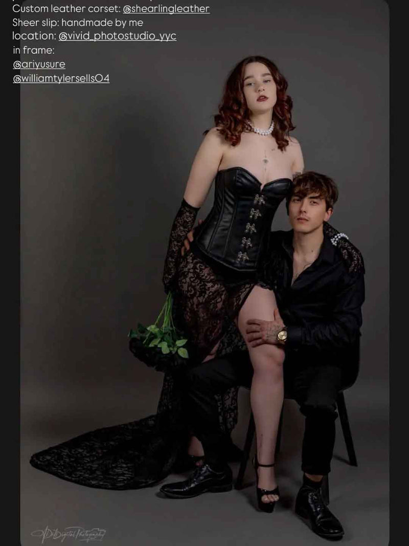 A custom black leather corset and sheer black lace slip set modeled by a woman and a man in a dark studio setting. The woman wears the corset with the lace slip layered underneath, holding a green floral arrangement, while the man sits beside her in a matching black outfit.