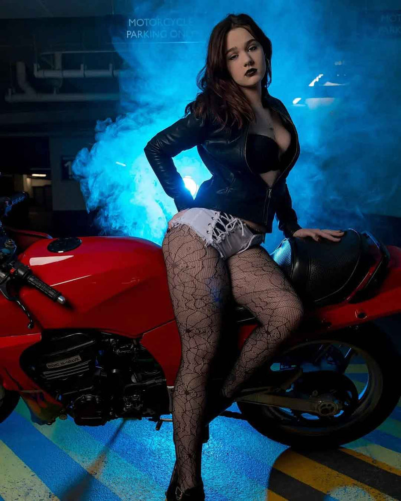 A woman posing on a red motorcycle in a smoky garage setting with blue lighting, wearing a black leather jacket, black lace-up bikini bottom, and black patterned fishnet stockings.
