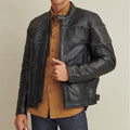 A man wearing a black leather rider jacket with a quilted design and covered front zipper, paired with a brown shirt and blue jeans.